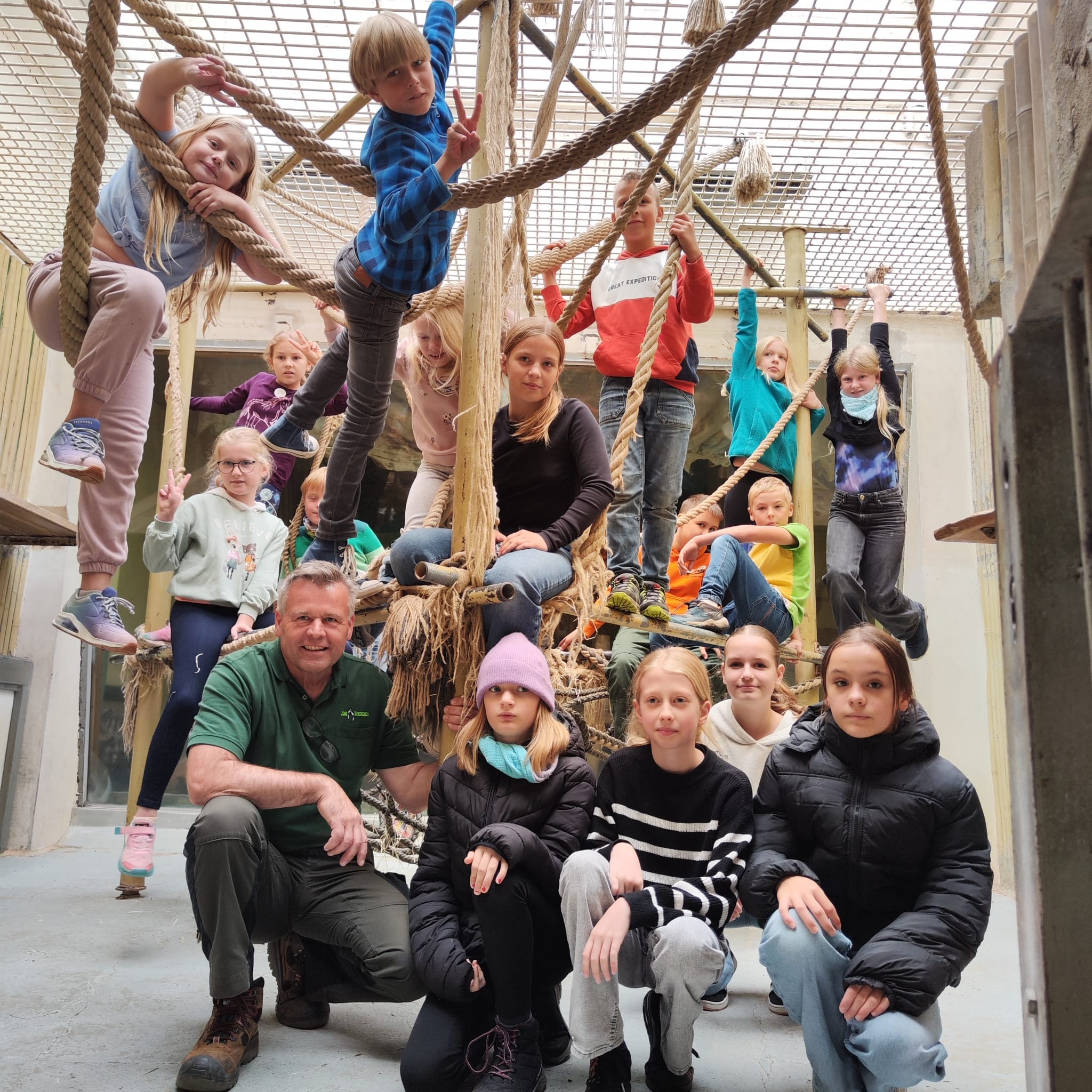 Erwachsener in grünem Polo mit Kindergruppe auf einem Klettergerüst aus Seilen und Holzstangen in einem Gebäude.