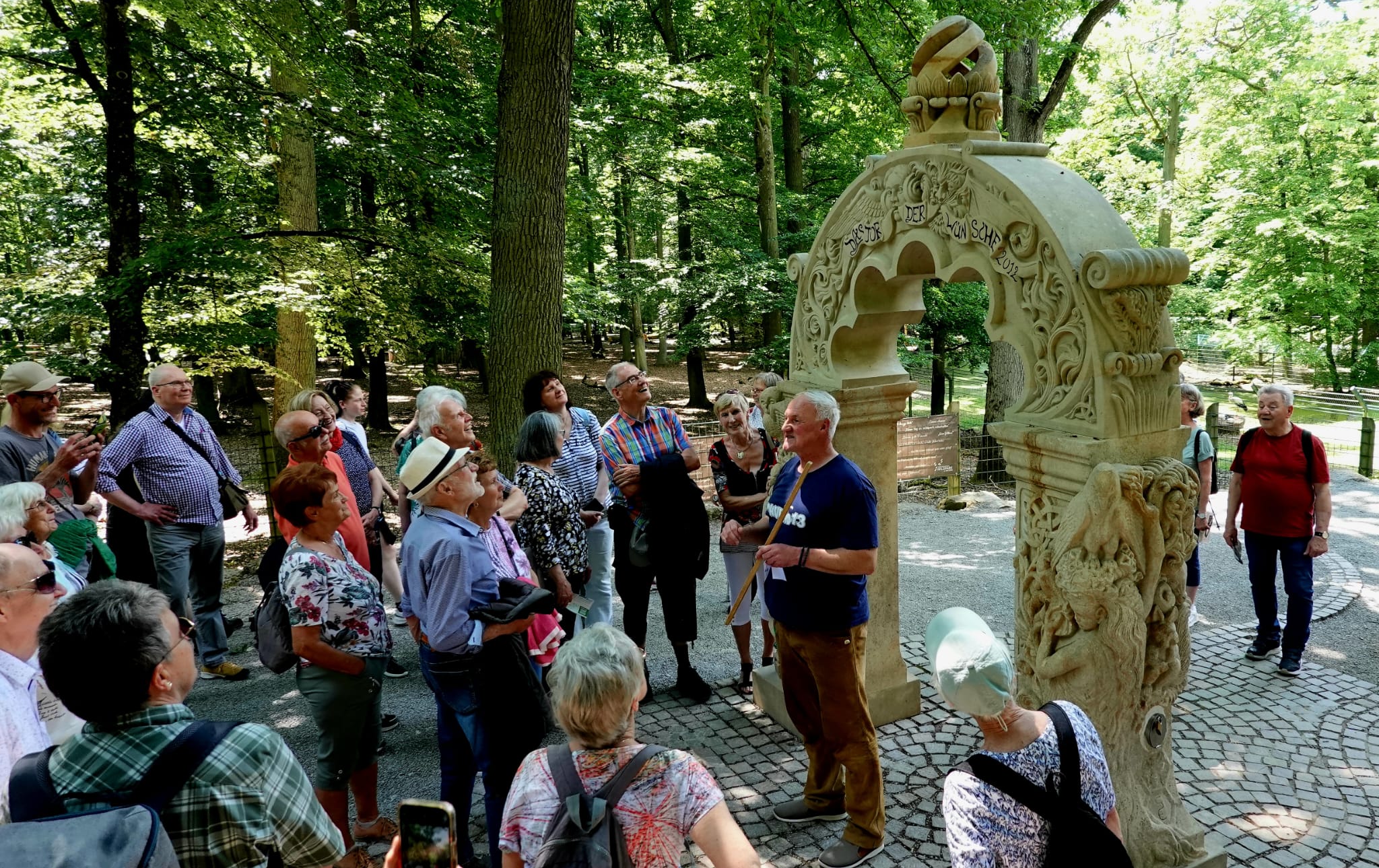 Reisegruppe betrachtet einen verzierten Sandstein-Brunnen im Wald, ein Mann mit Stock erklärt die Skulptur.