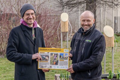 Zwei Männer halten gemeinsam ein Informationsschild mit der Aufschrift „Wandler im Sonnenlicht" in einem Garten.