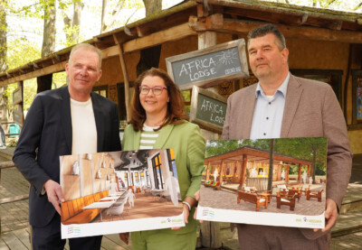 Drei Personen halten Architekturpläne vor einer Africa Lodge, umgeben von Bäumen.